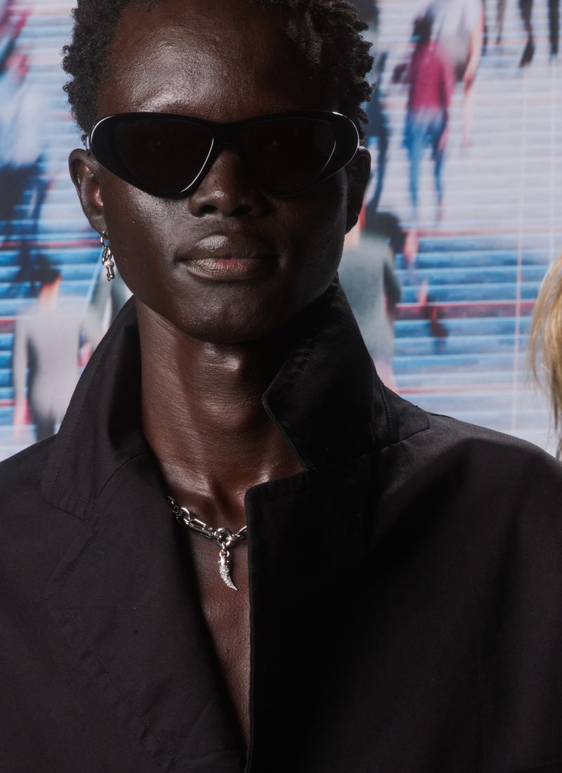 Close-up of a dark-skinned adult male model in sunglasses and a black coat, posing against a blurred backdrop with silhouetted figures.
