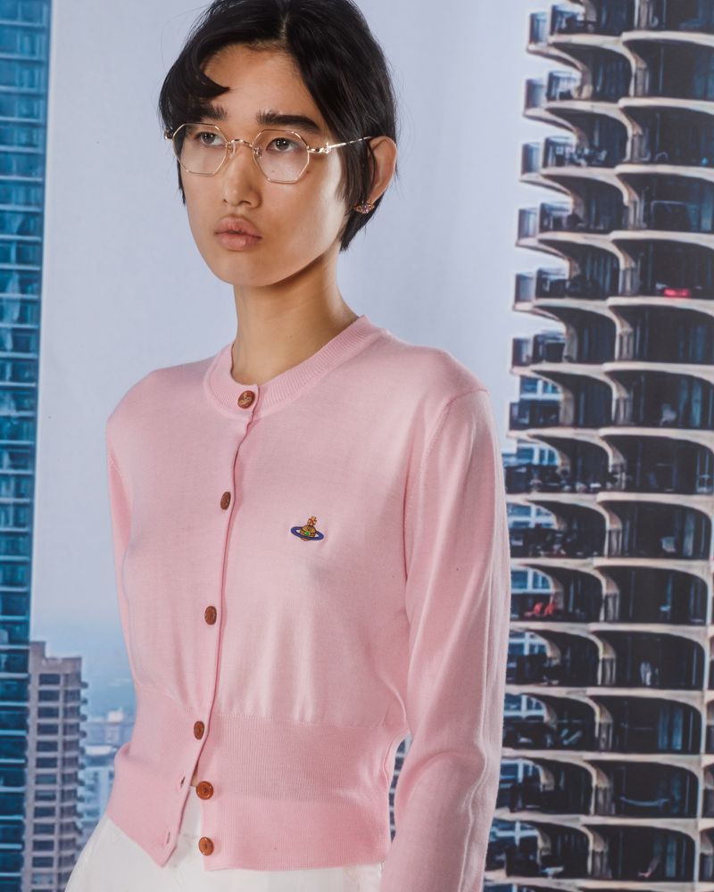 Woman wearing a pink cardigan and gold glasses stands against a backdrop of modern buildings on a bright day indoors.