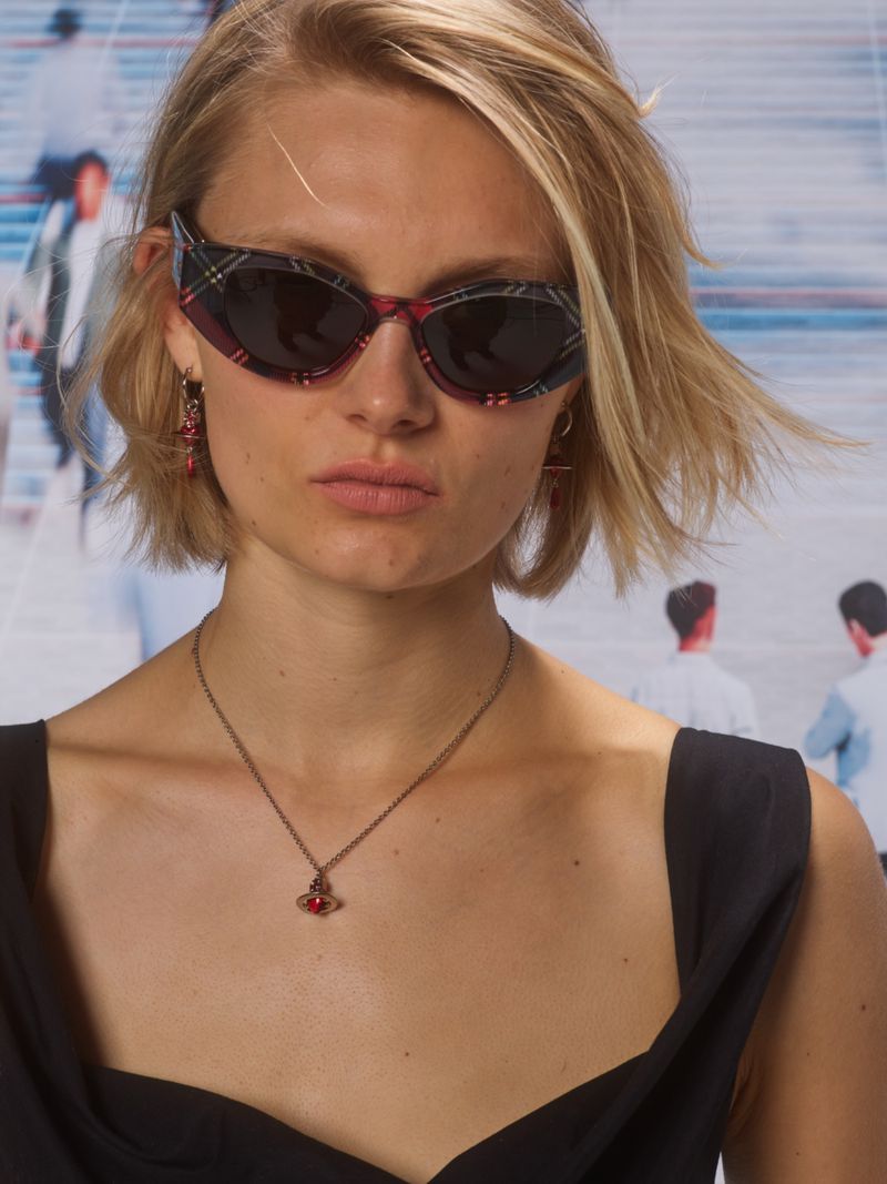 Close-up of a blonde woman wearing sunglasses with a red tartan pattern, along with a pendant necklace and earrings against a blurred background.