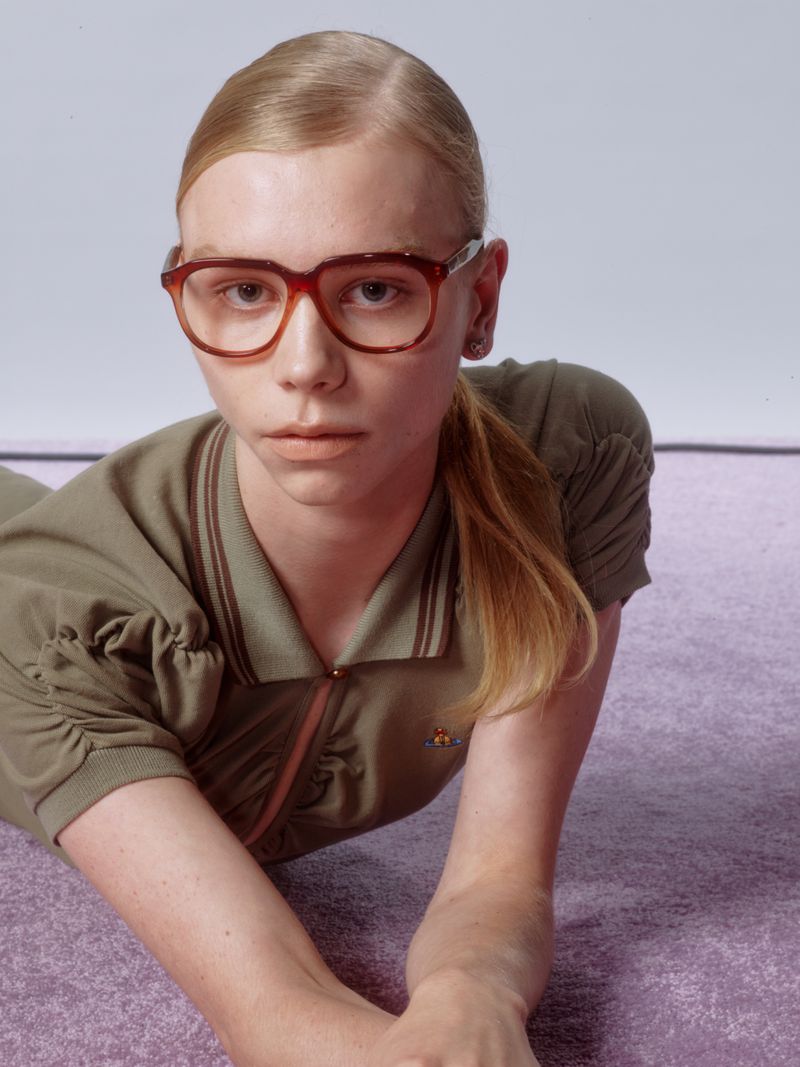 Woman with blonde hair poses indoors wearing an olive-green polo dress and brown-framed glasses, laying on a textured purple surface.
