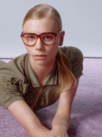 Woman with blonde hair poses indoors wearing an olive-green polo dress and brown-framed glasses, laying on a textured purple surface.