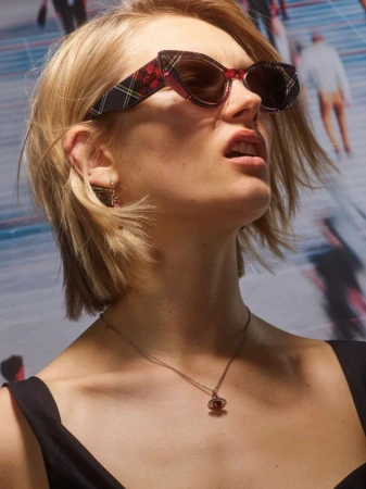 Close-up portrait of a woman wearing plaid cat-eye sunglasses and a necklace against a blurred urban street backdrop.