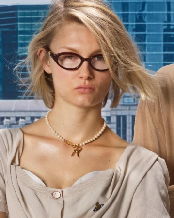 Close-up portrait of a woman wearing designer cat-eye glasses, a pearl necklace, and beige top against an urban background.
