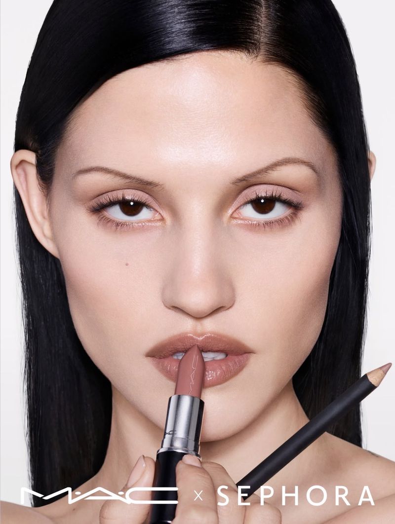 Close-up of a woman with black hair applying nude lipstick, promoting MAC x Sephora beauty collaboration, on a white background.