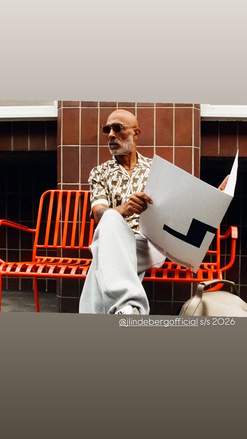 Stylish man with sunglasses sitting on an orange bench holding a poster in front of a brick wall, wearing a patterned shirt.