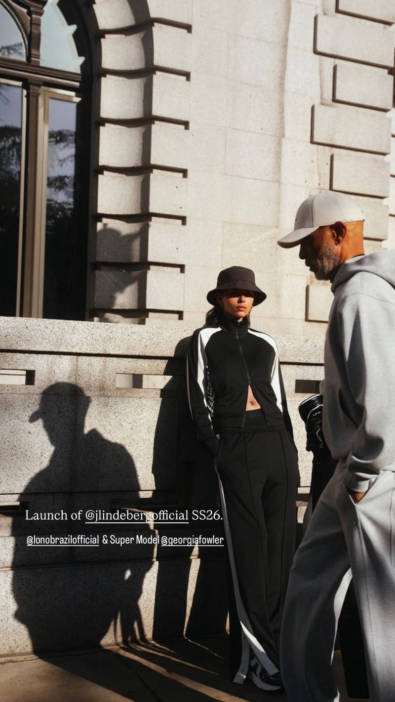 Two fashion models in grey and black tracksuits pose against an exterior wall with a window. The image features a mix of natural and shadow lighting.