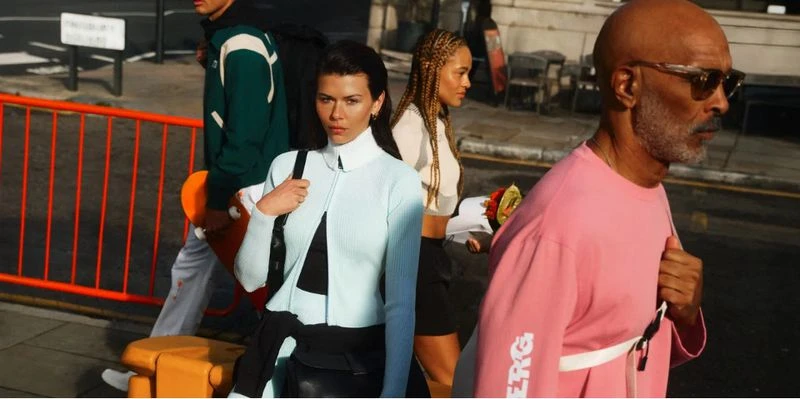 Four pedestrians on a sunny urban street: a woman in light blue, a man in pink, and others, create a vibrant street style fashion moment.