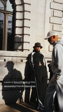Model Georgia Fowler in black tracksuit leaning against a stone wall with Lono Brazil walking past in a light grey hoodie.