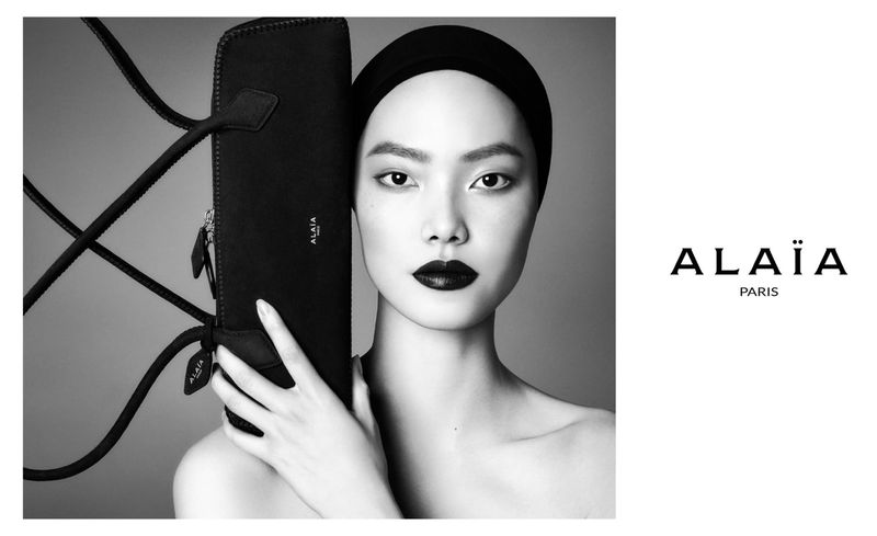 Monochrome portrait of an Asian woman holding a black handbag with the brand logo; close-up shot highlighting beauty and fashion.