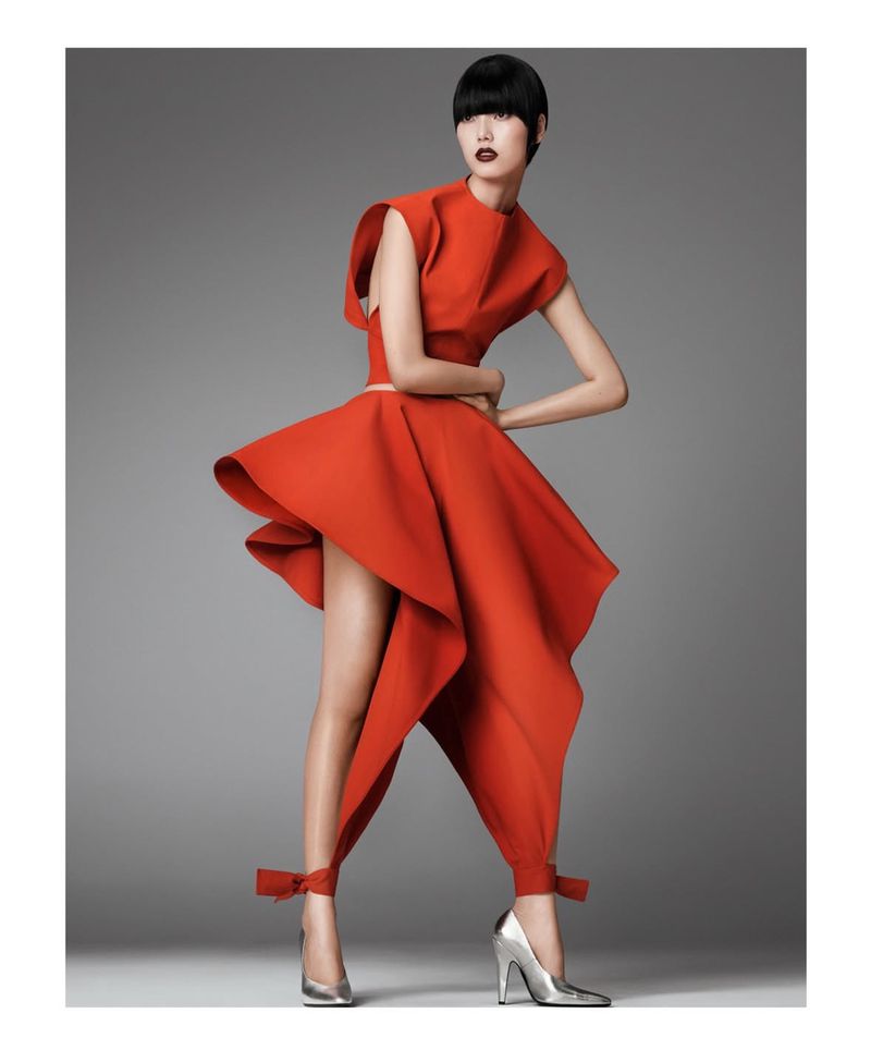 Fashion shot of a female model in a bright red couture dress with silver heels, posing in a studio with a light gray backdrop.