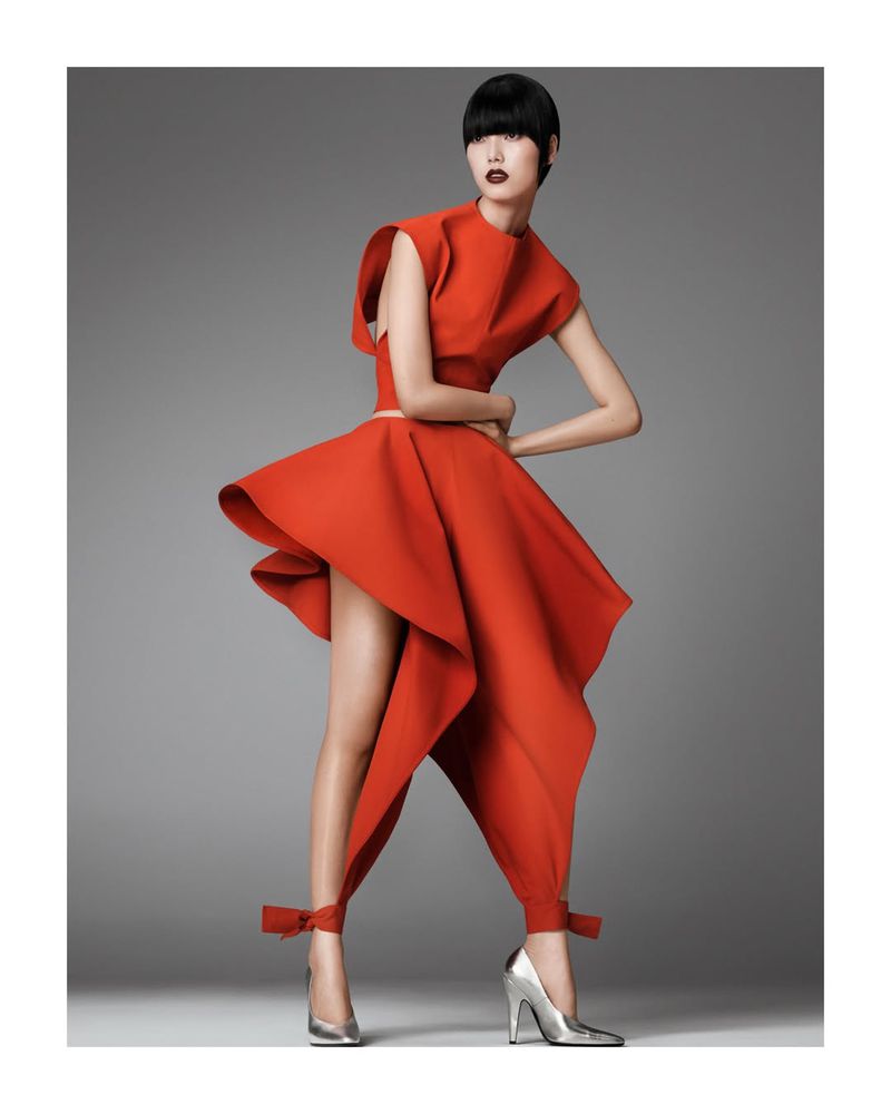 Elegant Asian woman in a bright red sculptural dress with silver high heels posing in a fashion studio with a neutral gray background.