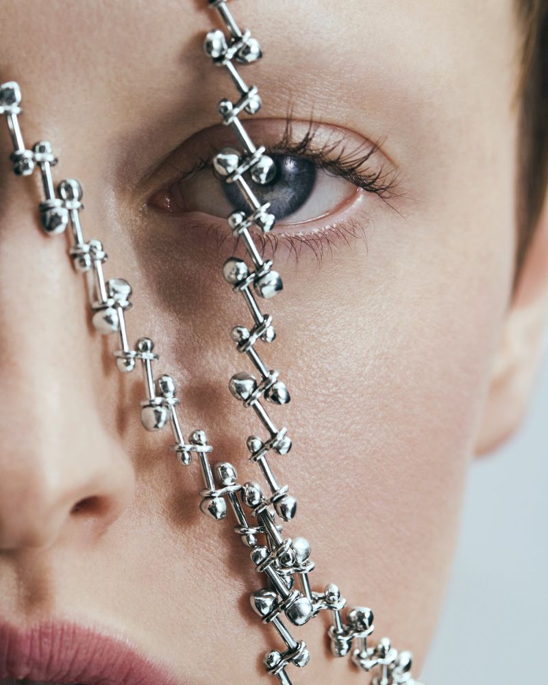Close-up of a woman's face with a silver linked chain accessory draped across her eye and face, showcasing a modern and minimalist aesthetic.