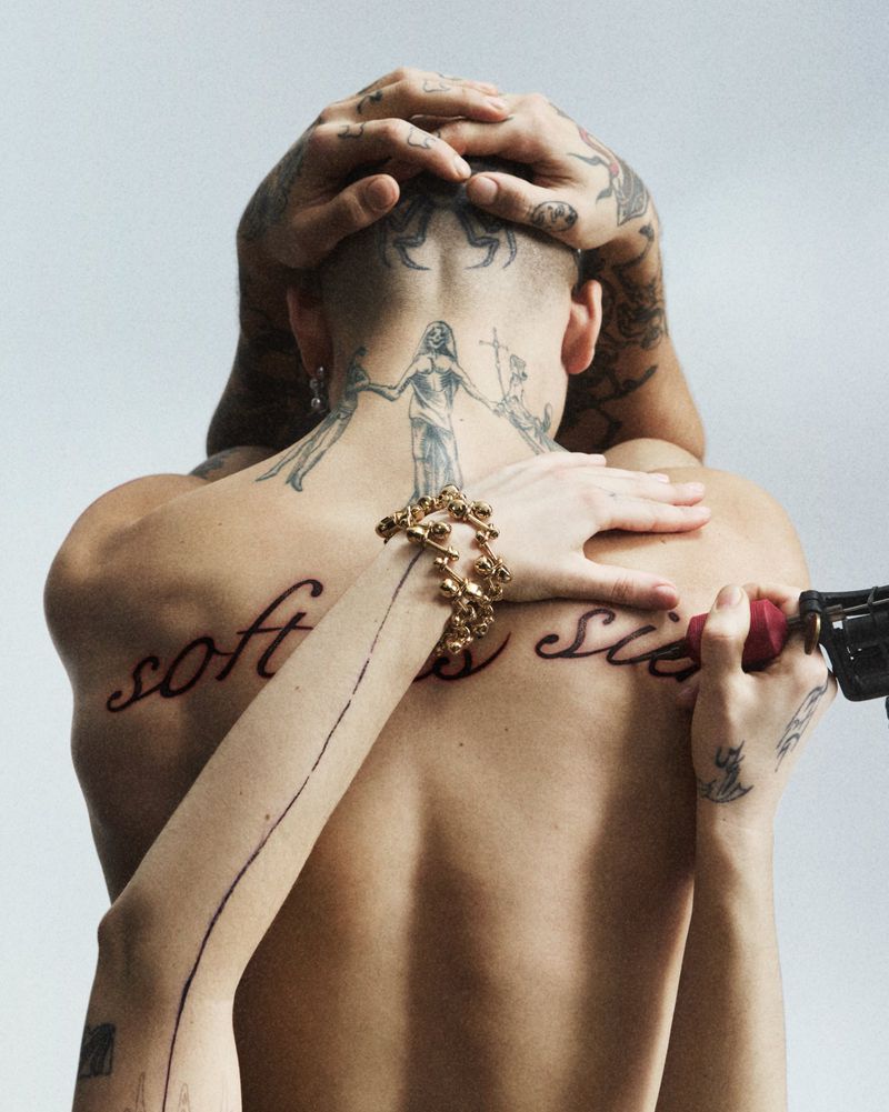 Back view of a heavily tattooed person getting a tattoo on their back. Hands are visible. They are wearing gold jewelry.
