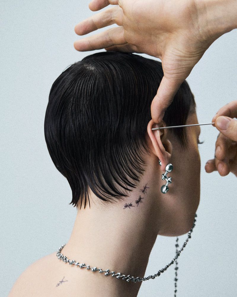An adult woman with short dark hair has a silver necklace and earrings. A hand pierces her ear with a needle against a light background.