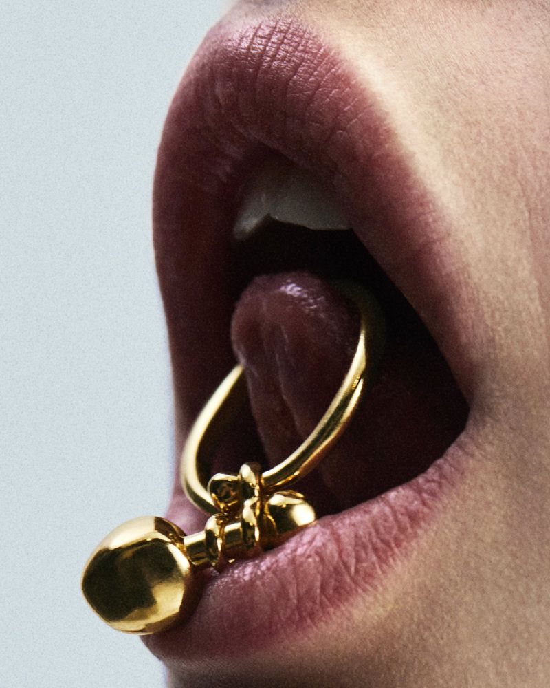 Close-up studio shot of a person's lips and tongue with a gold barbell piercing. The gold contrasts against the skin and light background.