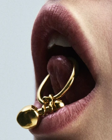 Close-up studio shot of a person's lips and tongue with a gold barbell piercing. The gold contrasts against the skin and light background.
