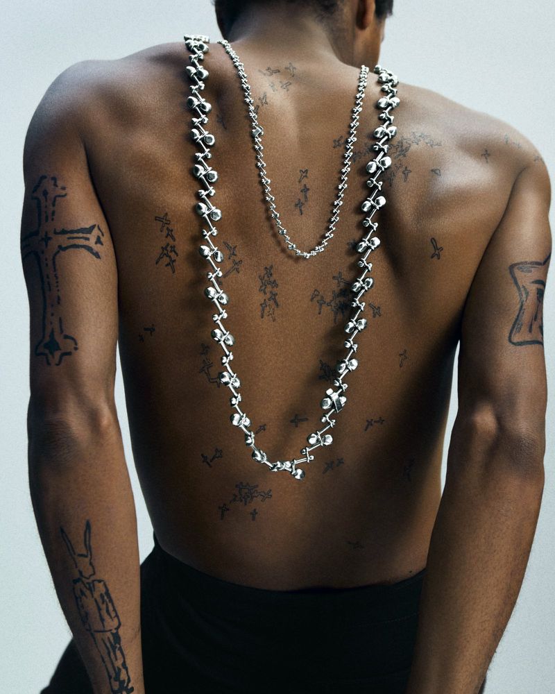 Upper torso of a dark-skinned adult male with neck jewelry and tattoos, set against a clean, neutral backdrop in a portrait.