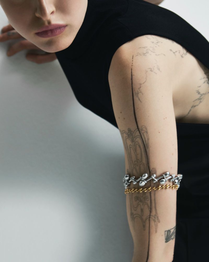 Close-up of a female model with tattoos on her arm, wearing a black dress and two metal armbands against a white wall.