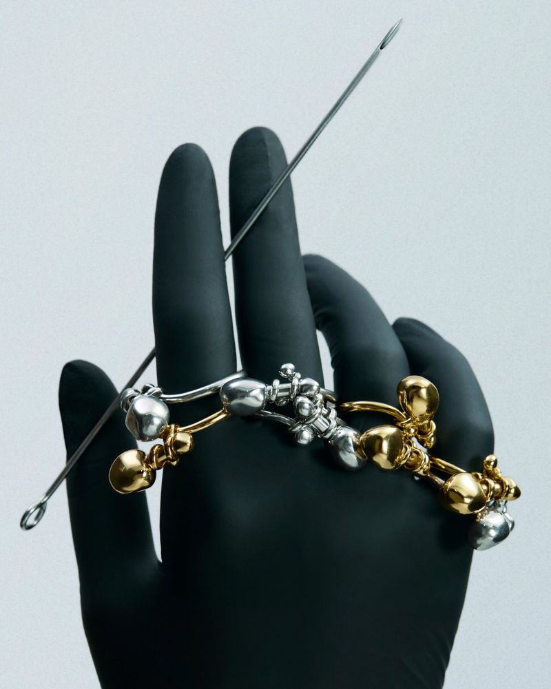 Stylized studio shot of a hand in a black glove holding silver and gold jewellery with a metal tattoo needle, against a light gray background.