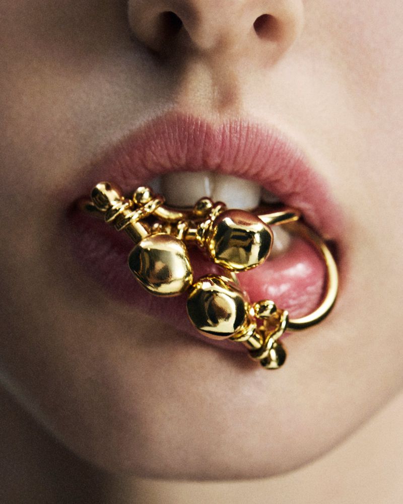 Close-up studio shot of a gold decorative item held in the mouth of an adult female, showing her lips, teeth, and tongue.
