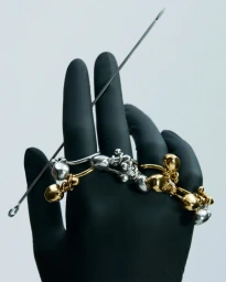 Gloved hand wearing gold and silver metallic jewelry pieces holding a long metal needle against a light background.