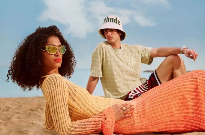 Two models pose on a sunny beach: a woman with gold sunglasses and a man in a bucket hat, showcasing fashion apparel and accessories.