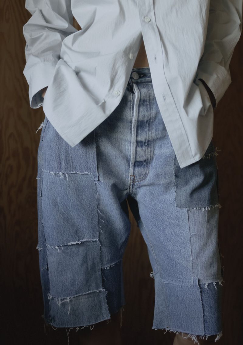 Close-up fashion shot of a person wearing patched denim shorts and a white button-down shirt against a wood-paneled wall.