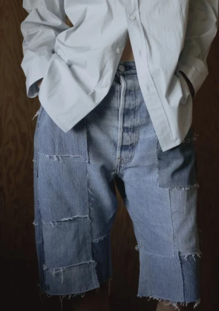 Close-up fashion shot of a person wearing patched denim shorts and a white button-down shirt against a wood-paneled wall.