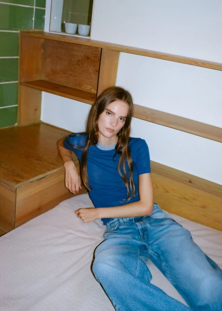 A young adult woman lies on a bed, wearing a blue t-shirt and jeans, indoors, with natural lighting.