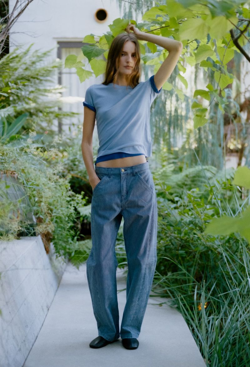 A woman poses in a blue T-shirt and blue jeans in a garden setting, offering a natural and relaxed aesthetic. Soft light.