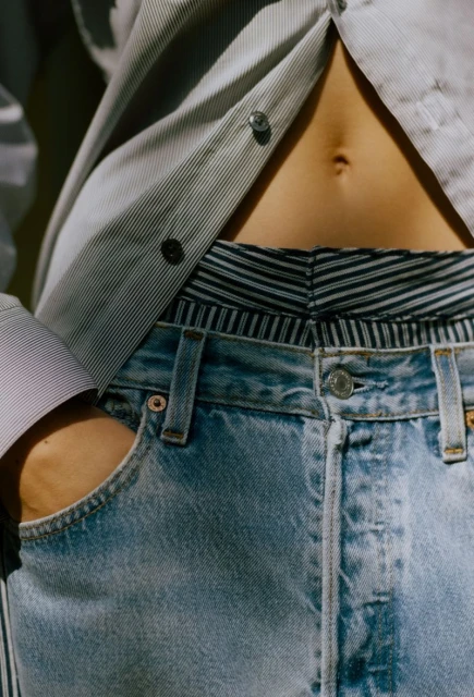 Fashion shot of a woman wearing blue jeans, and a pinstripe button-down shirt, showing midriff, in a vertical, close-up composition.