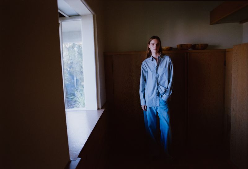 Adult woman in a light blue shirt and jeans standing inside of a building with light coming in from a window in daytime.