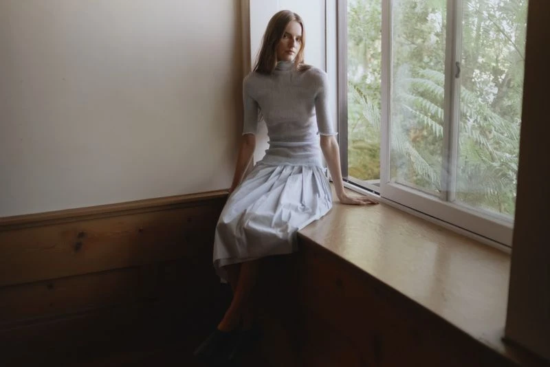 An adult woman poses sitting in front of a window inside a home interior. She wears a light blue dress in a fashion editorial style.