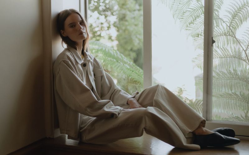 Adult female model in beige outfit poses sitting in a window, bathed in natural light with a lush green view beyond the glass.