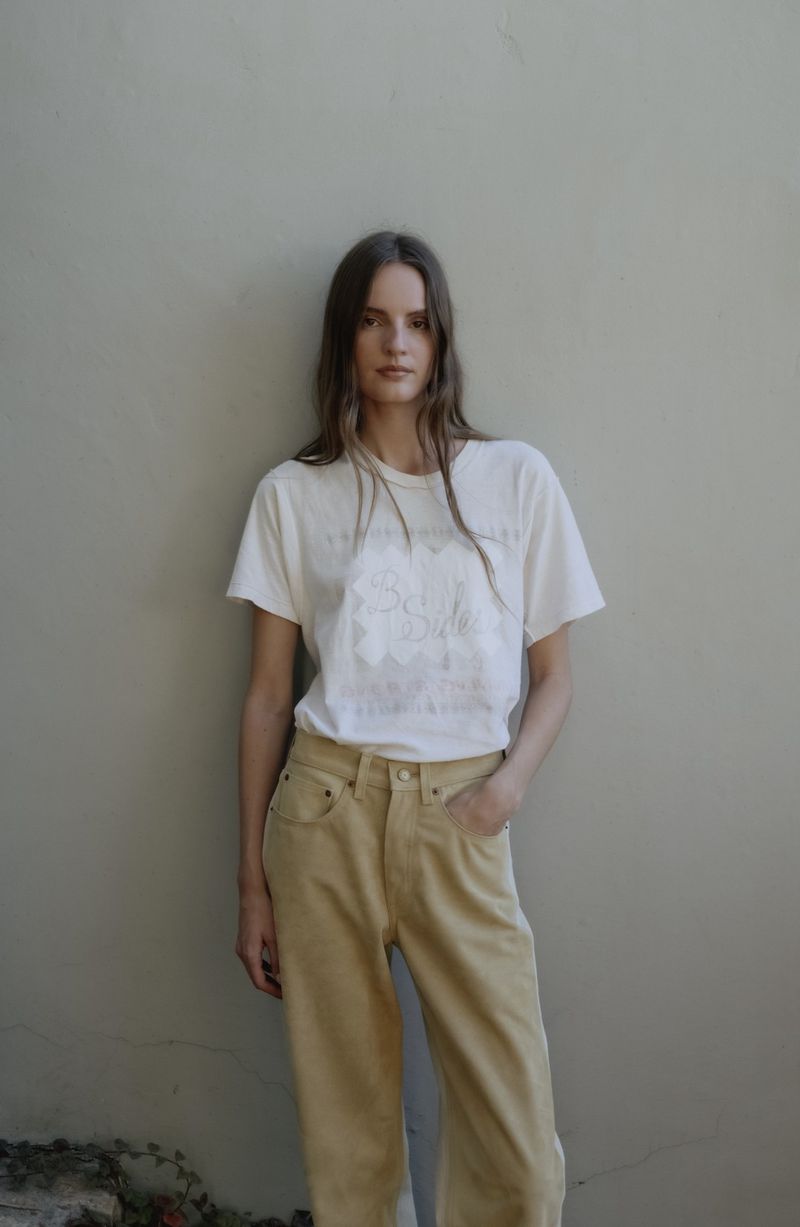 Model Tilda Lindstam in a white t-shirt and yellow jeans leaning against a neutral wall for B Sides Jeans S/S 26 campaign.