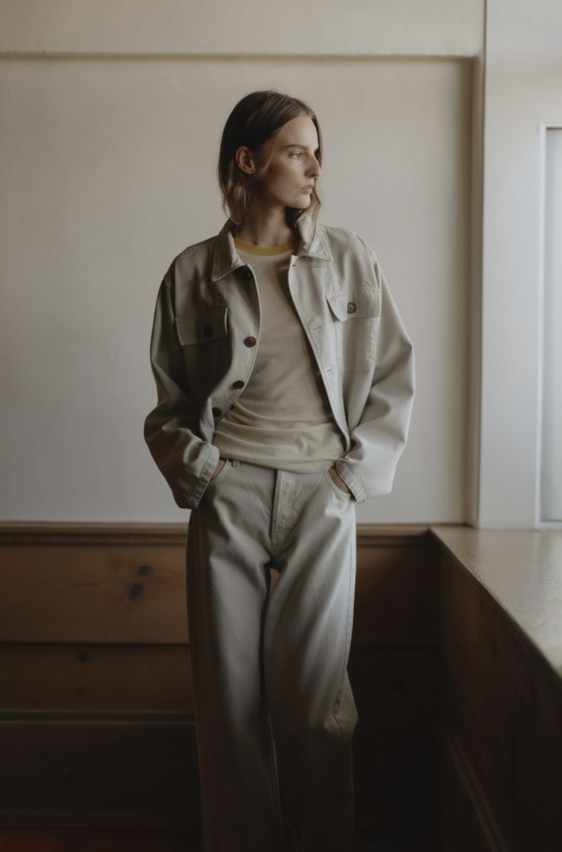 Indoor portrait of a female fashion model wearing matching beige jacket and pants, looking away from the camera near a window.