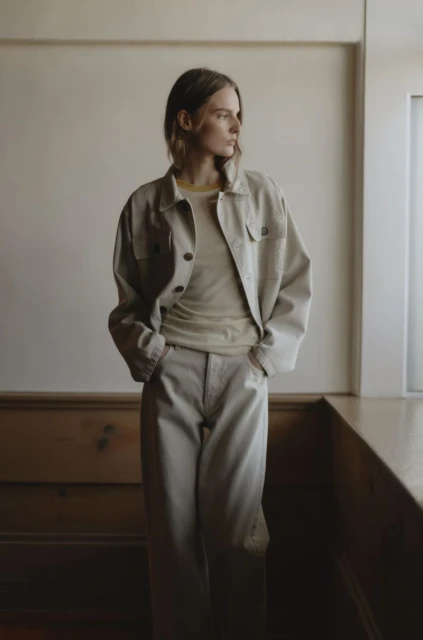 Indoor portrait of a female fashion model wearing matching beige jacket and pants, looking away from the camera near a window.