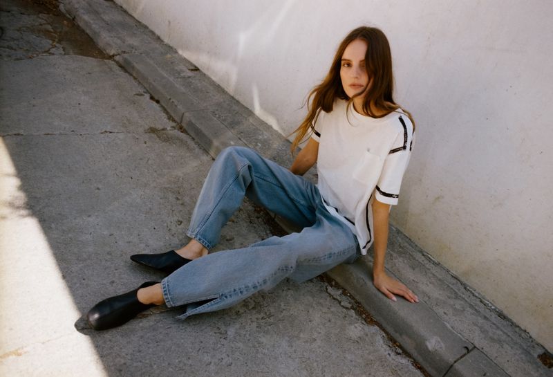 Model Tilda Lindstam seated on a concrete curb wearing a white t-shirt, blue jeans, and black shoes for B Sides Jeans.
