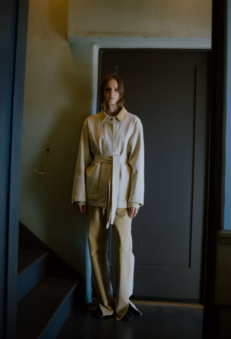A female model poses in a beige belted jacket and pants beside a dark door and staircase indoors.