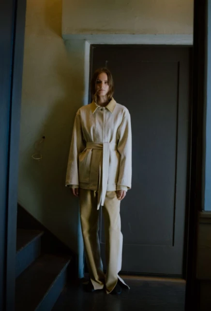 A female model poses in a beige belted jacket and pants beside a dark door and staircase indoors.