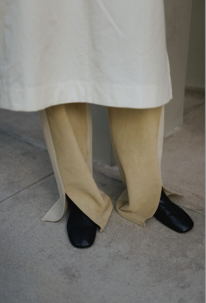 Person wearing an off-white coat and tan pants with black shoes stands on a light grey surface.