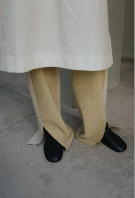 Person wearing an off-white coat and tan pants with black shoes stands on a light grey surface.
