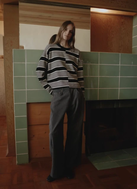 Adult woman in a striped shirt and wide-leg pants poses indoors by a tiled fireplace, with a mirror reflecting the natural light.