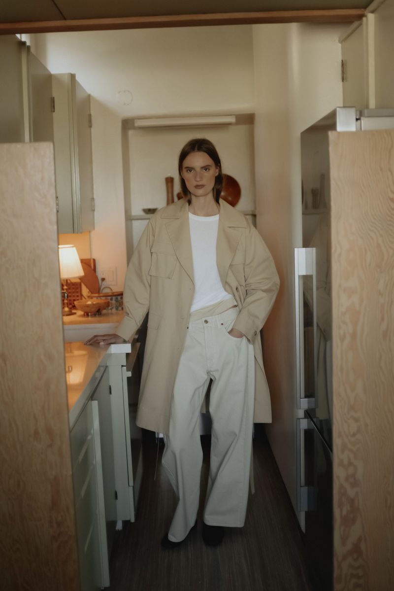 Fashionable adult female in a white T-shirt and beige trench coat poses in a narrow kitchen with neutral-toned cabinets and lighting.