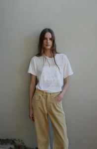 Model Tilda Lindstam in a white t-shirt and yellow jeans leaning against a neutral wall for B Sides Jeans S/S 26 campaign.