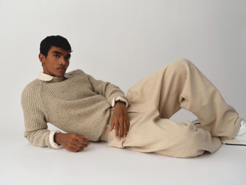 Man lounges in a beige ribbed sweater and wide-leg pants against a light backdrop, offering a relaxed yet stylish fashion statement.