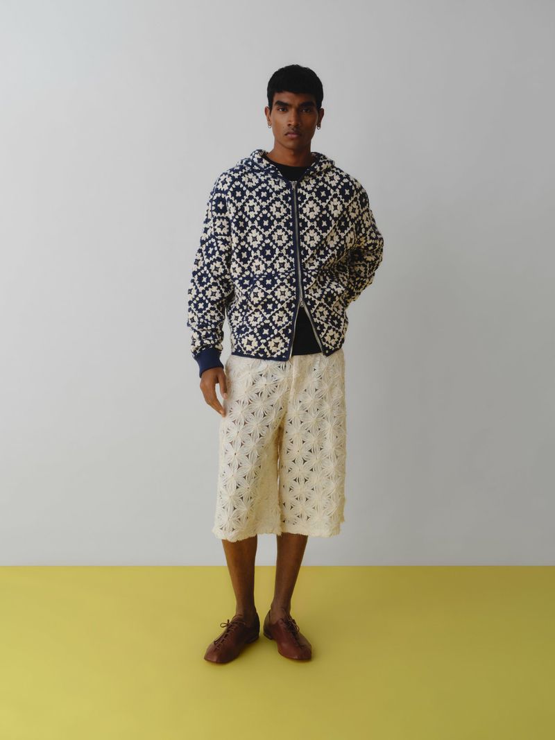 A full shot of a male model in a blue and white patterned zip-up jacket and off-white crochet shorts, posing against a light background.