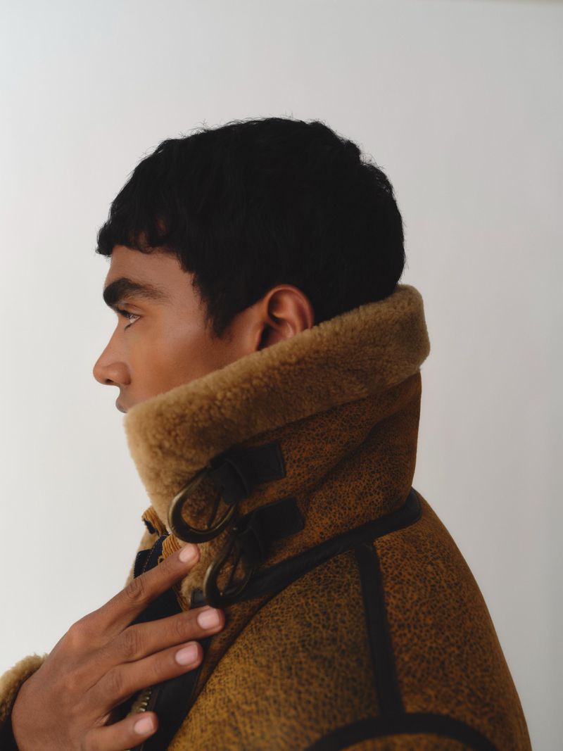 Close-up portrait of a man in a shearling jacket, side profile with his hand near the jacket collar, against a plain white background.