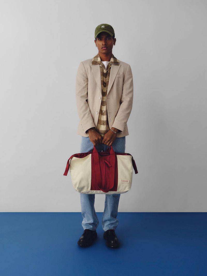 Model wearing a beige blazer and holding a large cream and red duffel bag against a light studio wall with a blue floor line.