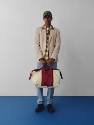Model wearing a beige blazer and holding a large cream and red duffel bag against a light studio wall with a blue floor line.